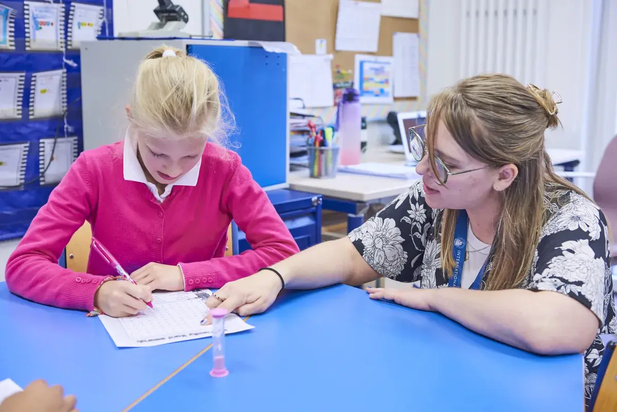 Teacher assisting a young student with writing at a classroom desk