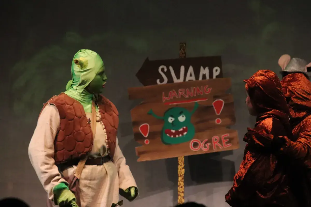 Student actors performing Shrek on stage next to a Swamp warning sign prop